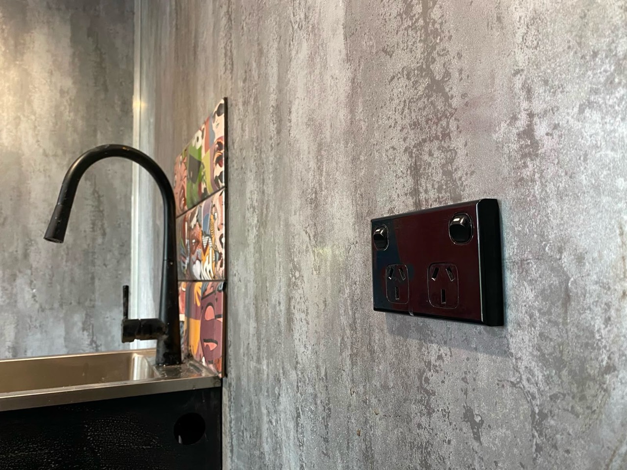 Black double power point fitted to a textured kitchen splashback beside a black tap